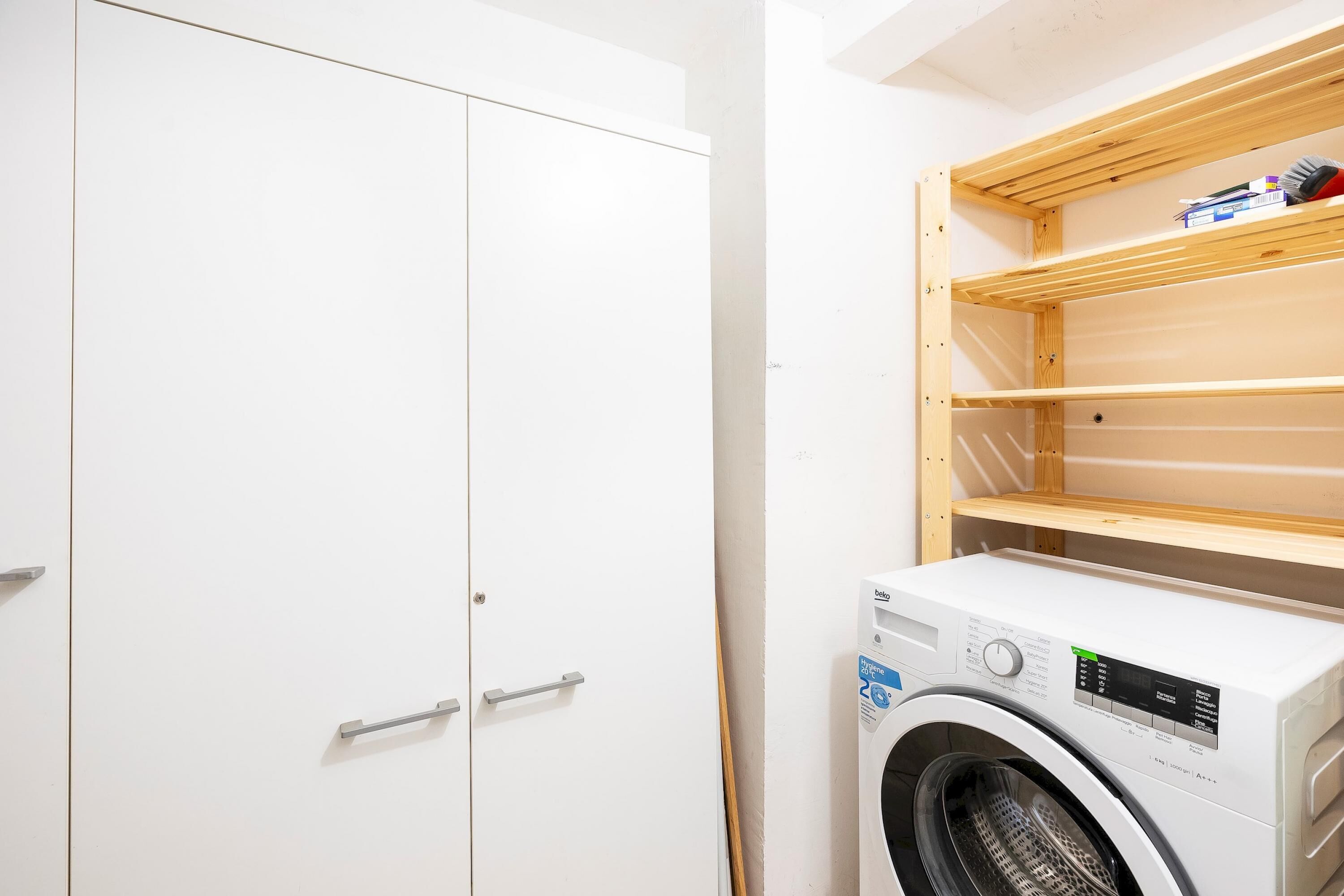 A laundry room with a white washing machine, wooden shelves, and a white wardrobe.