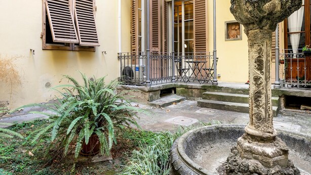 A courtyard with a stone fountain, a potted plant, and a building with wooden shutters and a balcony. - Venezia Double in Firenze (Florence)