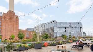 A rooftop terrace with outdoor seating, a view of a modern building, and a fire exit sign. - Battersea Power Station Flat With River Views (London)
