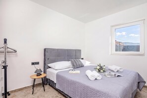 Un lit bien fait, avec une tête de lit grise, une petite table de chevet sur laquelle se trouve une lampe, et une fenêtre donnant sur un immeuble.