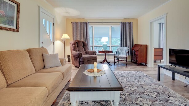 Un salon avec un canapé beige, une table basse en bois foncé sur laquelle est posée une bougie, un fauteuil marron, une petite table avec une lampe et un téléviseur sur un meuble.