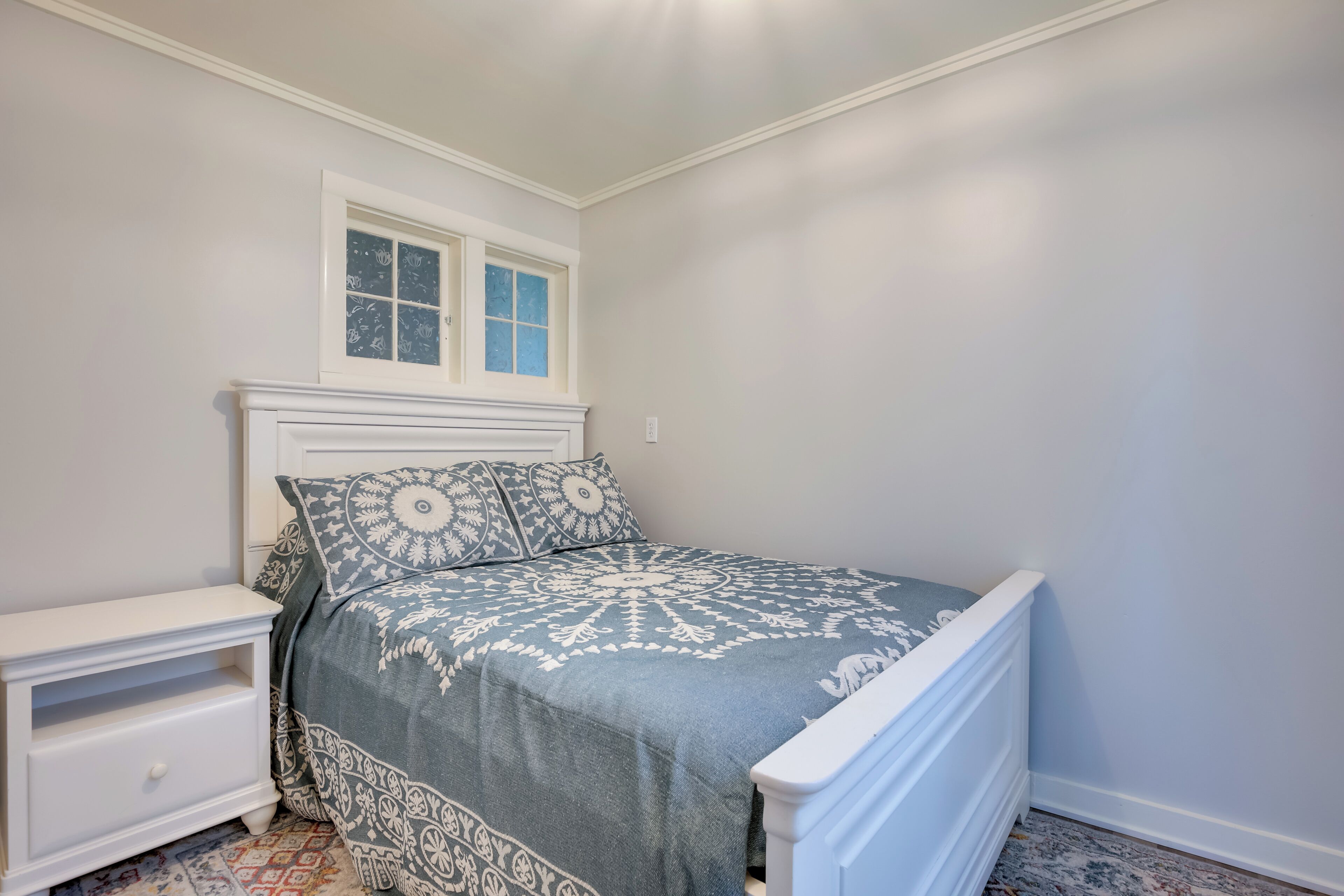 Quarto com cama, mesa de cabeceira, janela e tapete estampado.