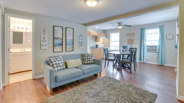 A living room with a sofa, a rug, a dining area with chairs and a table, a kitchen with cabinets and appliances, and a ceiling fan.
