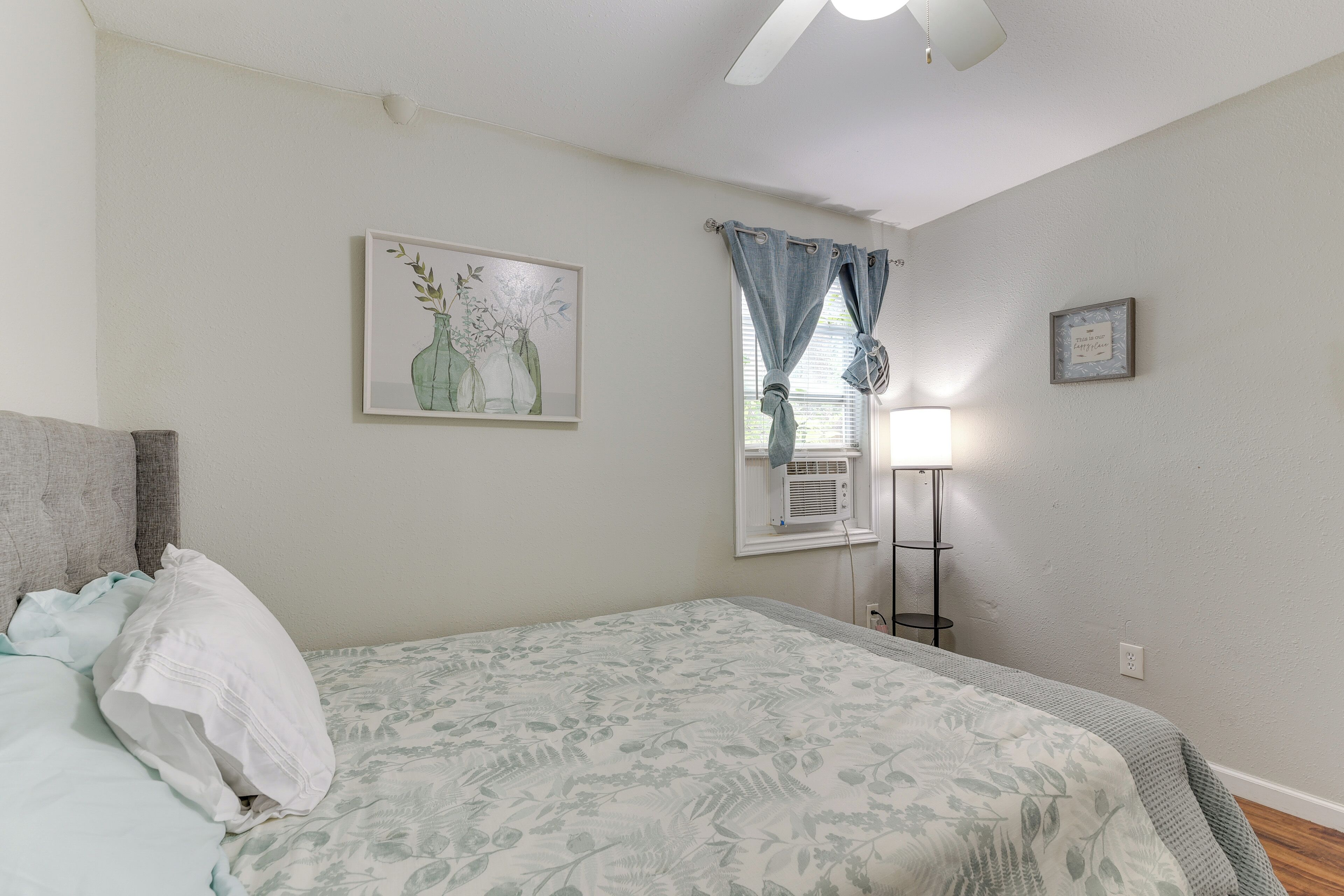 A bedroom with a bed, a nightstand, a lamp, a window with curtains, and a framed picture on the wall.