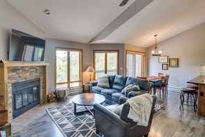 Uma sala de estar espaçosa com lareira, uma televisão de tela plana grande fixada acima, um sofá de couro, uma área de jantar com mesa e cadeiras, e janelas amplas que proporcionam luz natural.