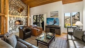 A cozy living room with a stone fireplace, leather sofas, a flat-screen TV, and a large window with a mountain view. - 98 Ridgepoint 4 Bedroom Condo by RedAwning (Avon)