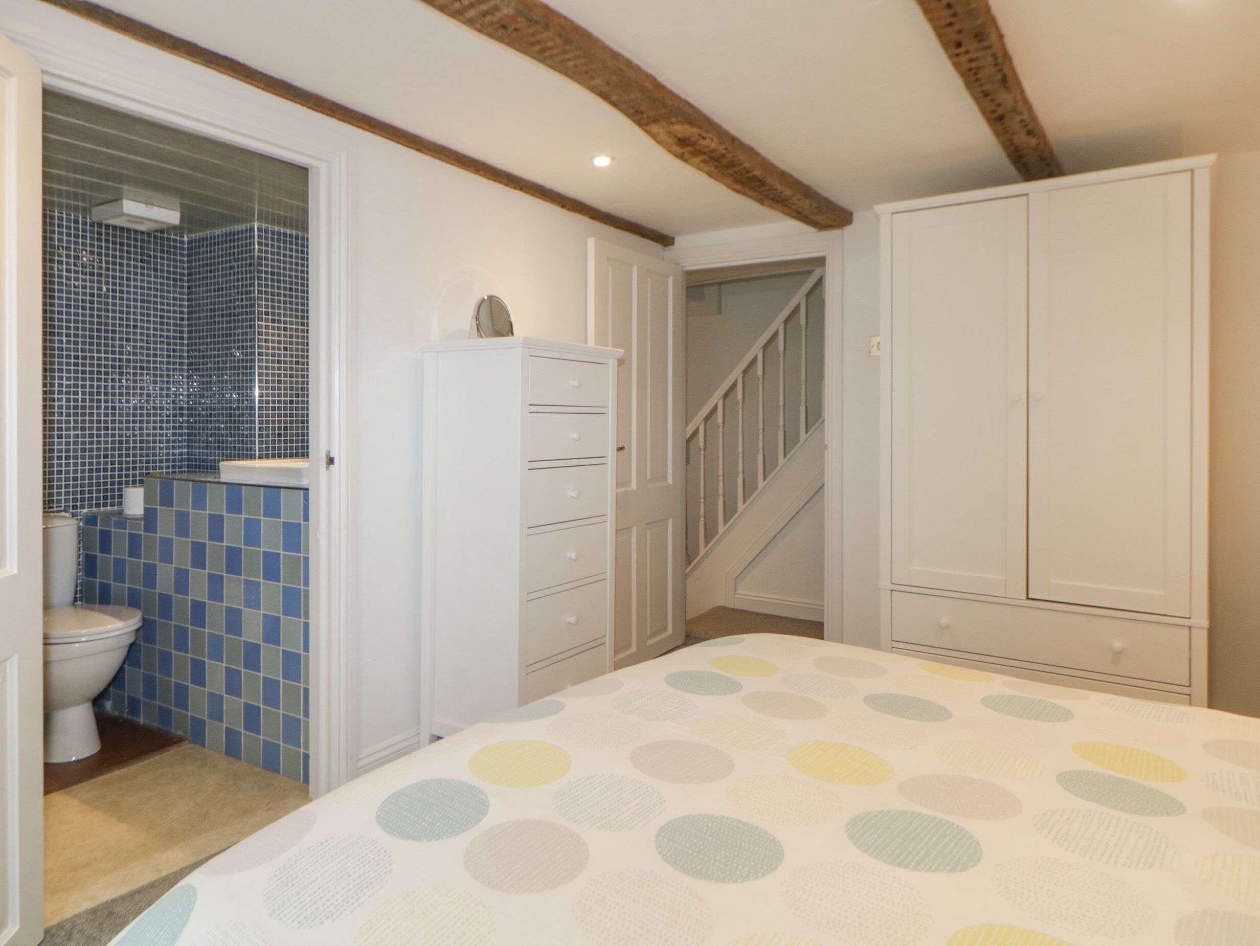 A bedroom with a bed, a wardrobe, a chest of drawers, a staircase, and a bathroom with a toilet and blue-tiled walls.