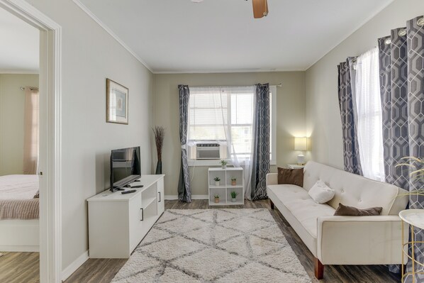 A compact living room with a sofa, a TV, a small white cabinet, a radiator, and a window with curtains. - ~ 1 Mi to the Strand: Central Unit in Galveston! (Galveston)