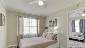 A bedroom with a bed, a ceiling fan, a window with curtains, a bedside table with a lamp, and a view into another room with a couch. - ~ 1 Mi to the Strand: Central Unit in Galveston! (Galveston)