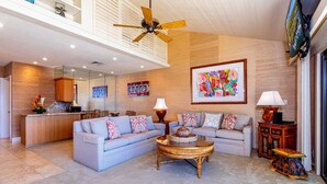 A spacious living area with a high ceiling, a ceiling fan, a large painting, and a modern kitchen in the background.
