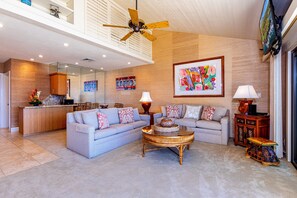 Un vaste espace de vie doté d’un haut plafond, d’un ventilateur de plafond, d’un grand tableau et d’une cuisine moderne en arrière-plan.