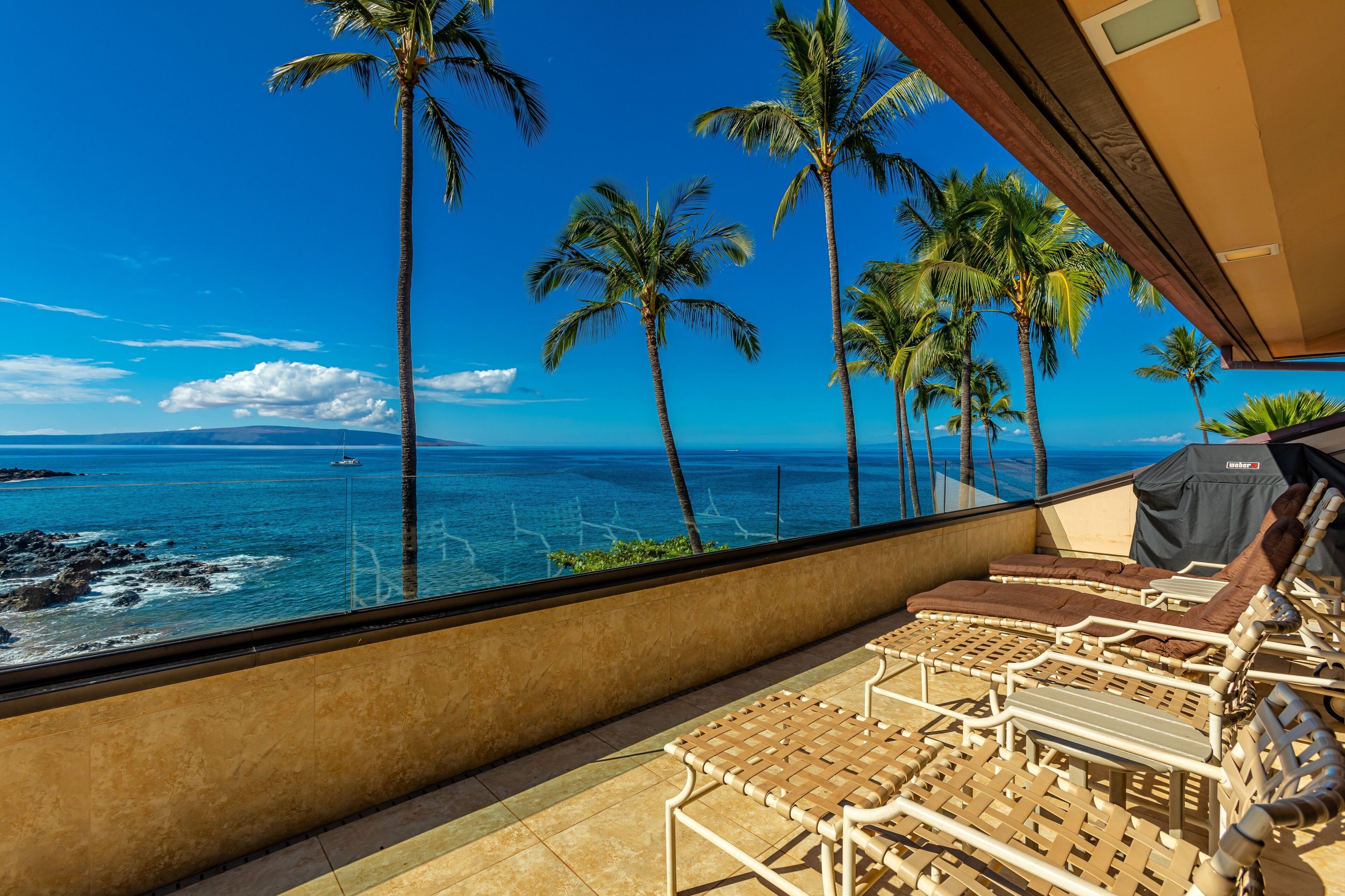 A balcony with lounge chairs, a view of the ocean, and palm trees.