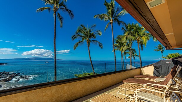 A balcony with lounge chairs, a view of the ocean, and palm trees.
