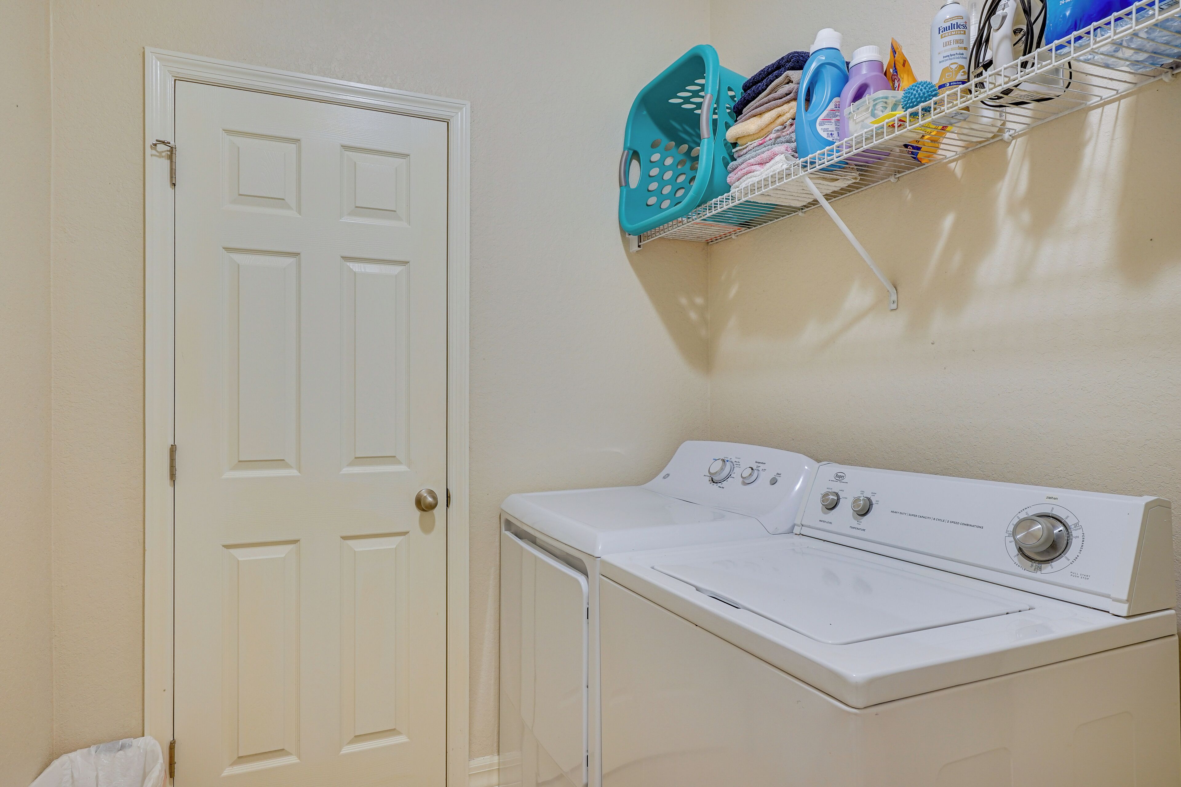 A laundry room with a white door, a stacked washer and dryer, and a shelf with cleaning supplies.