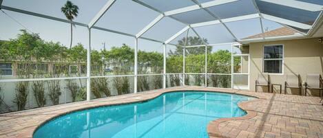 A swimming pool with a curved edge, surrounded by a brick patio and a covered area with a metal frame.