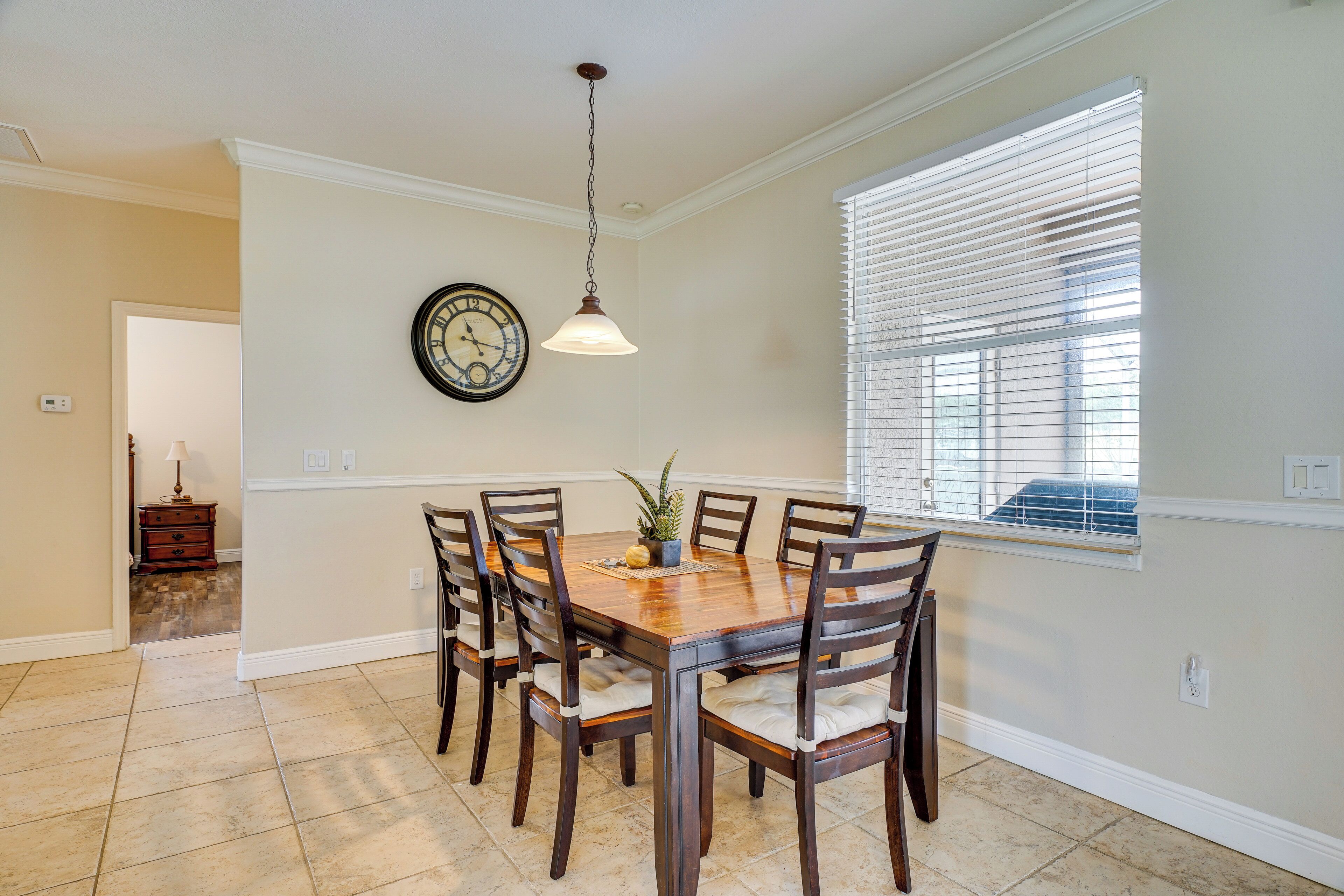 Comedor con mesa y sillas de madera, lámpara colgante, reloj en la pared y ventana con persianas.