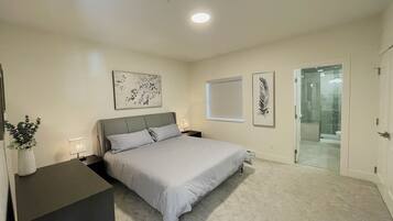Une chambre à coucher comprenant un lit, des tables de chevet, un bureau, une plante en pot et une porte donnant sur une salle de bain.