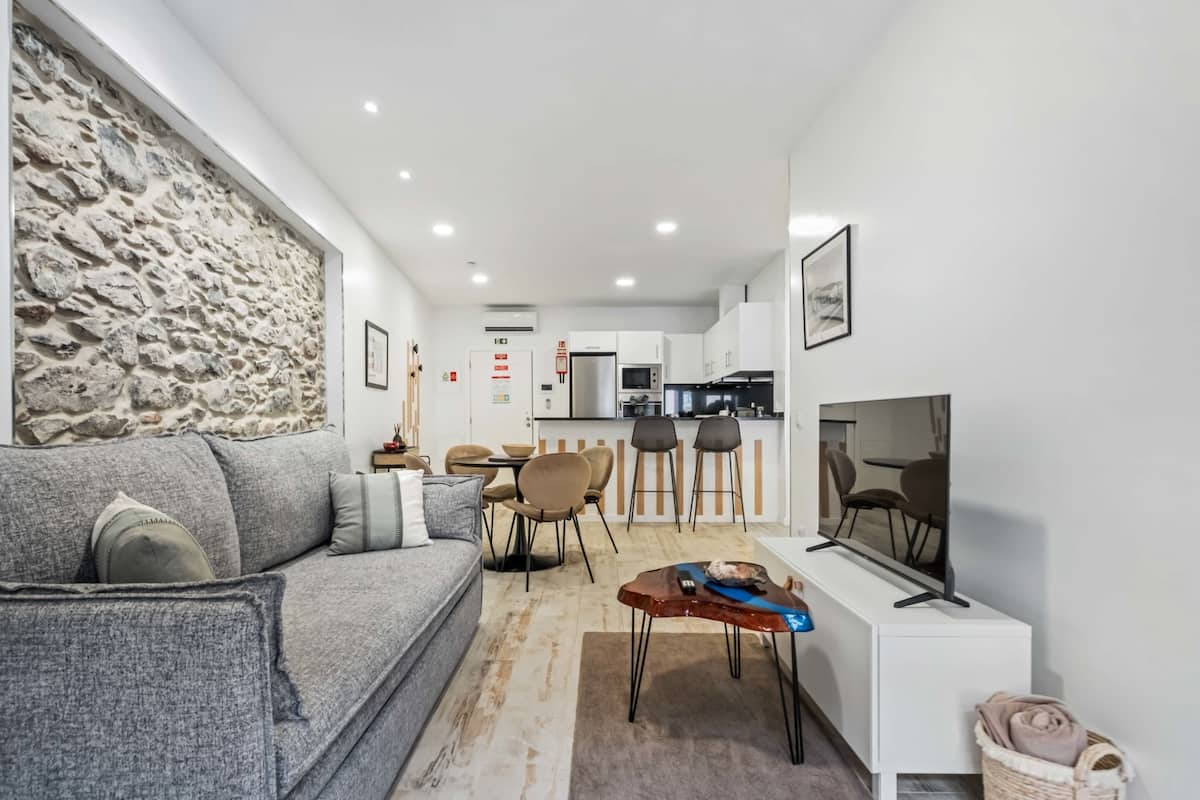 Un salón moderno con sofá gris, pared de piedra decorativa, zona de comedor con sillas, cocina equipada y televisor sobre un mueble blanco.