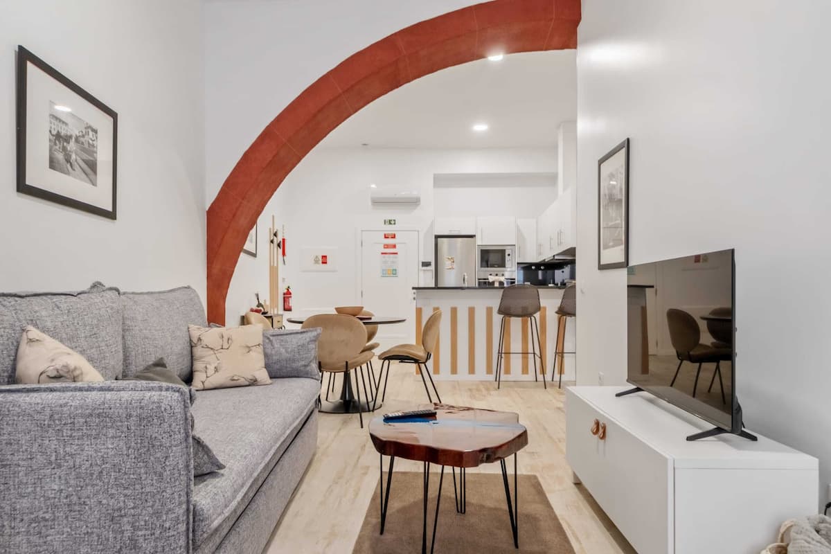 Una sala de estar moderna con un sofá gris, una mesa de centro y un televisor sobre un mueble blanco. Un arco de entrada da a una zona de cocina con un refrigerador, microondas y taburetes de bar.
