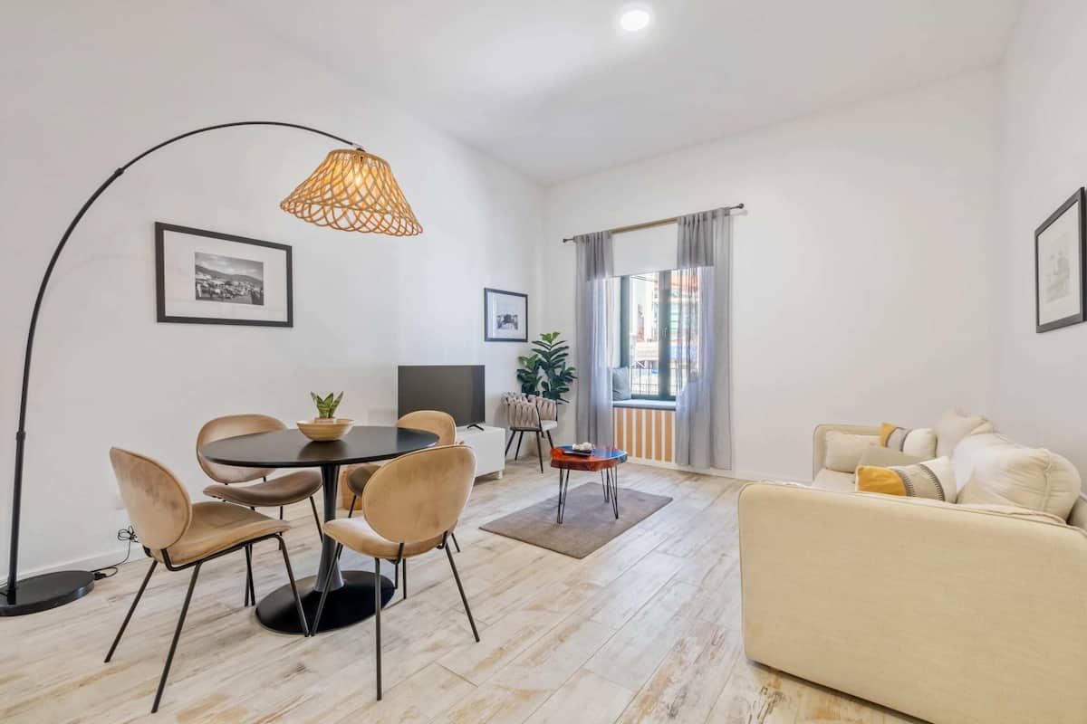 Una sala de estar moderna con una mesa de comedor redonda, un sofá color beige y un amplio ventanal con cortinas.