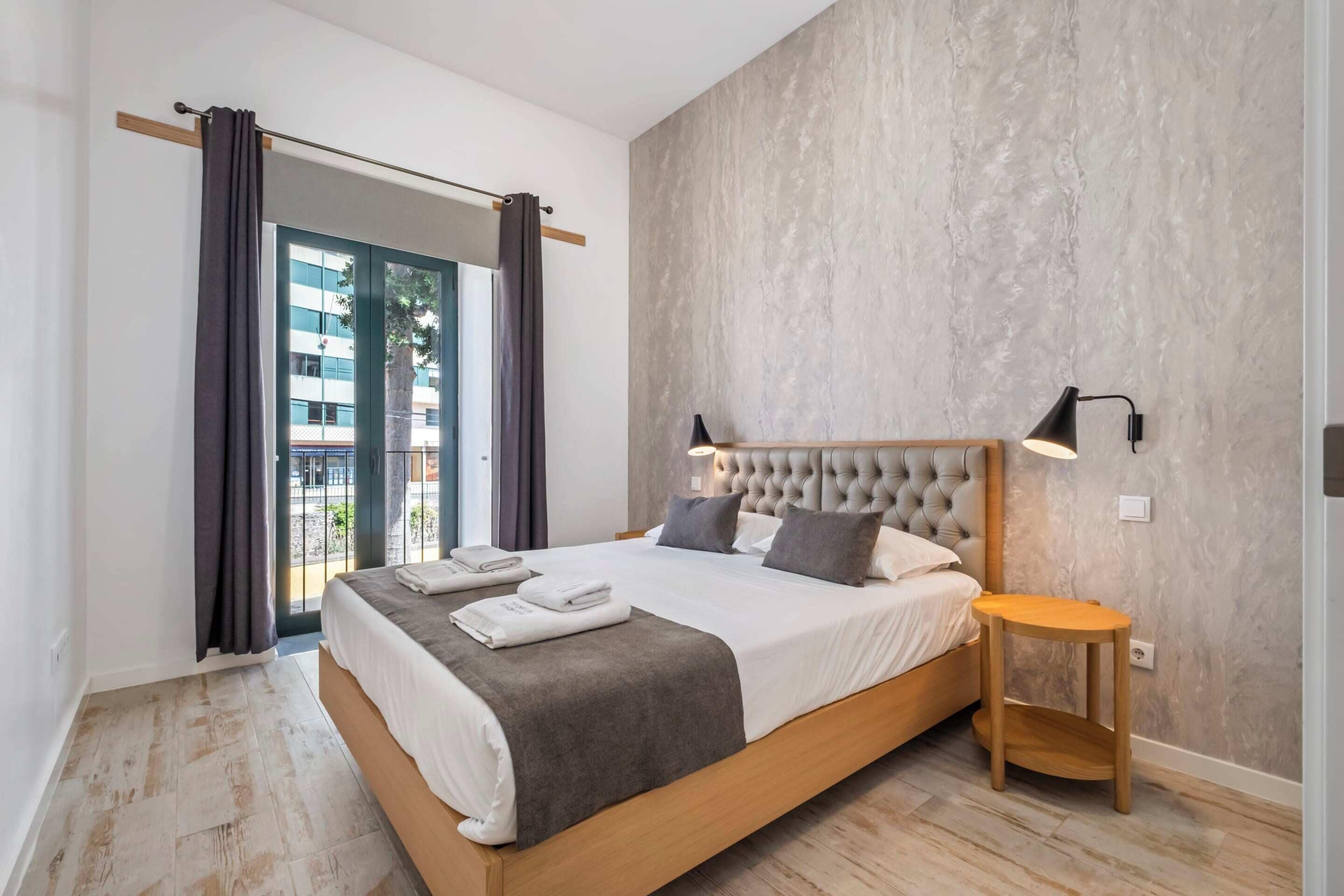 Ein modernes Schlafzimmer mit einem großen Bett, einem Holzkopfende, einem Nachttisch und Blick ins Freie durch eine Glastür.