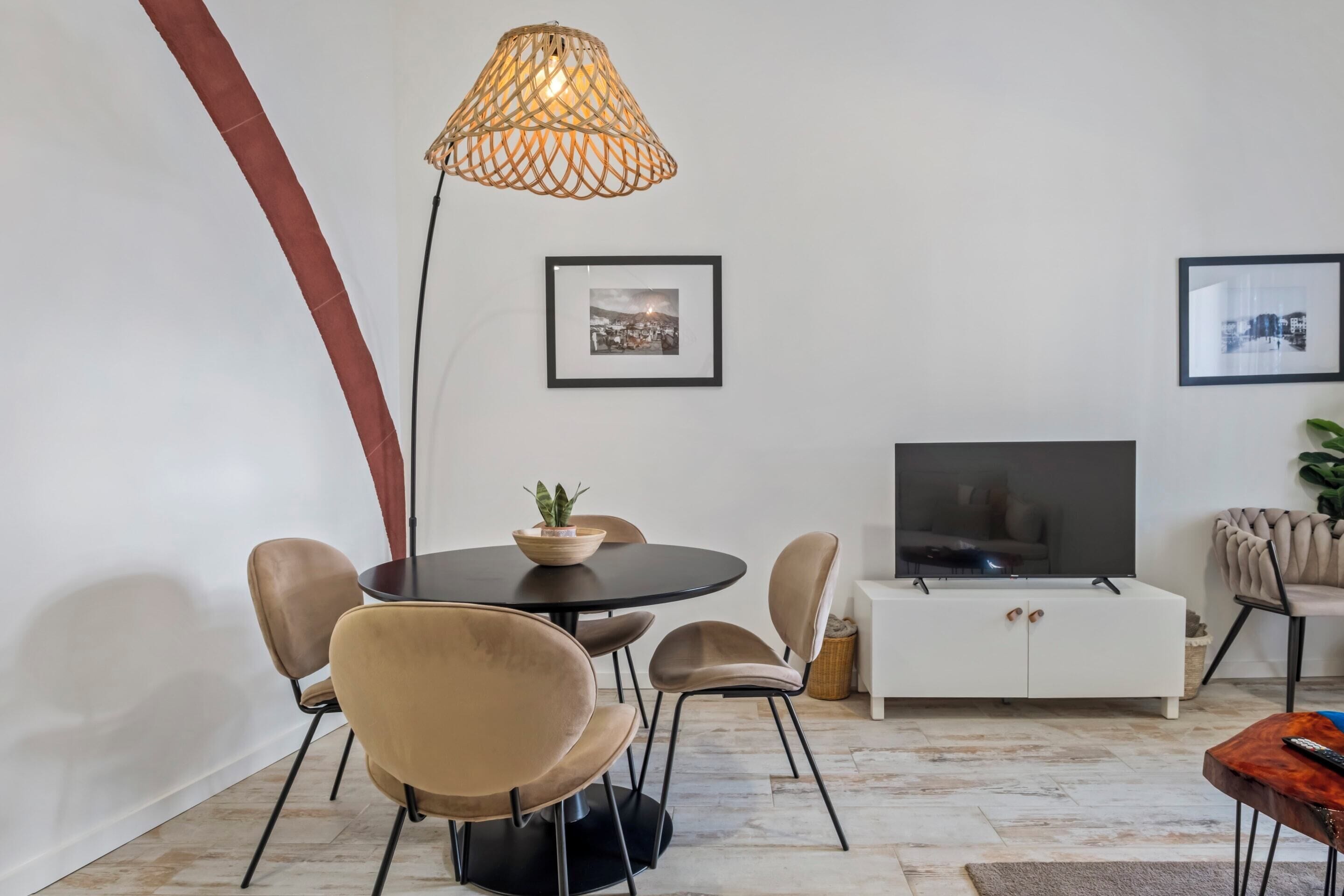 A modern living room with a round dining table, beige chairs, a TV on a white cabinet, and a hanging light fixture.
