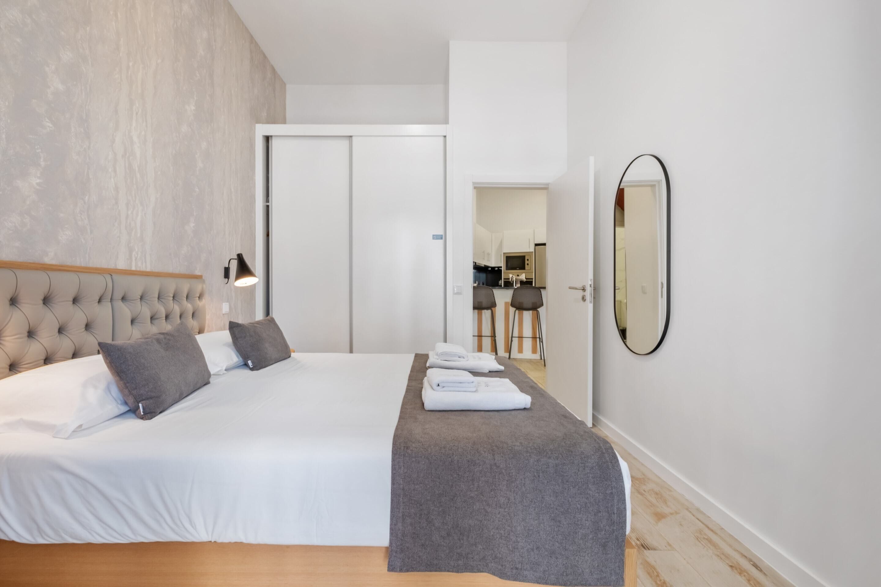 Una habitación de hotel moderna con una cama grande, un banco gris, un espejo montado en la pared y una puerta que da a una zona de cocina.