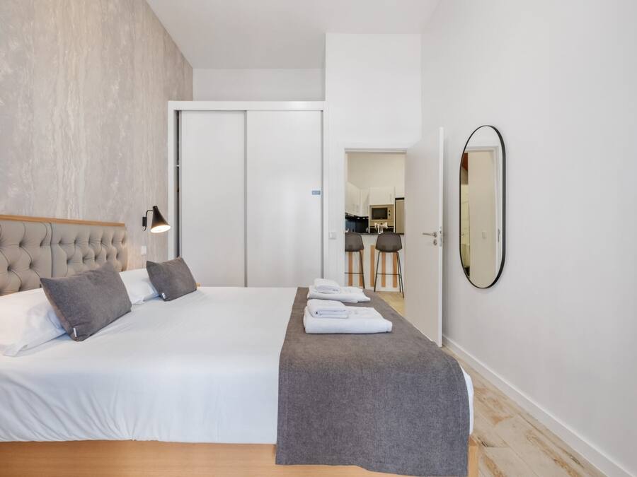 Una habitación de hotel moderna con una cama grande, un banco gris, un espejo montado en la pared y una puerta que da a una zona de cocina.