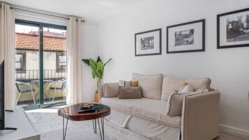 Ein modernes Wohnzimmer mit einer Couch, einem Couchtisch und einem großen Fenster mit Vorhängen. An der Wand hängen zwei gerahmte Bilder.