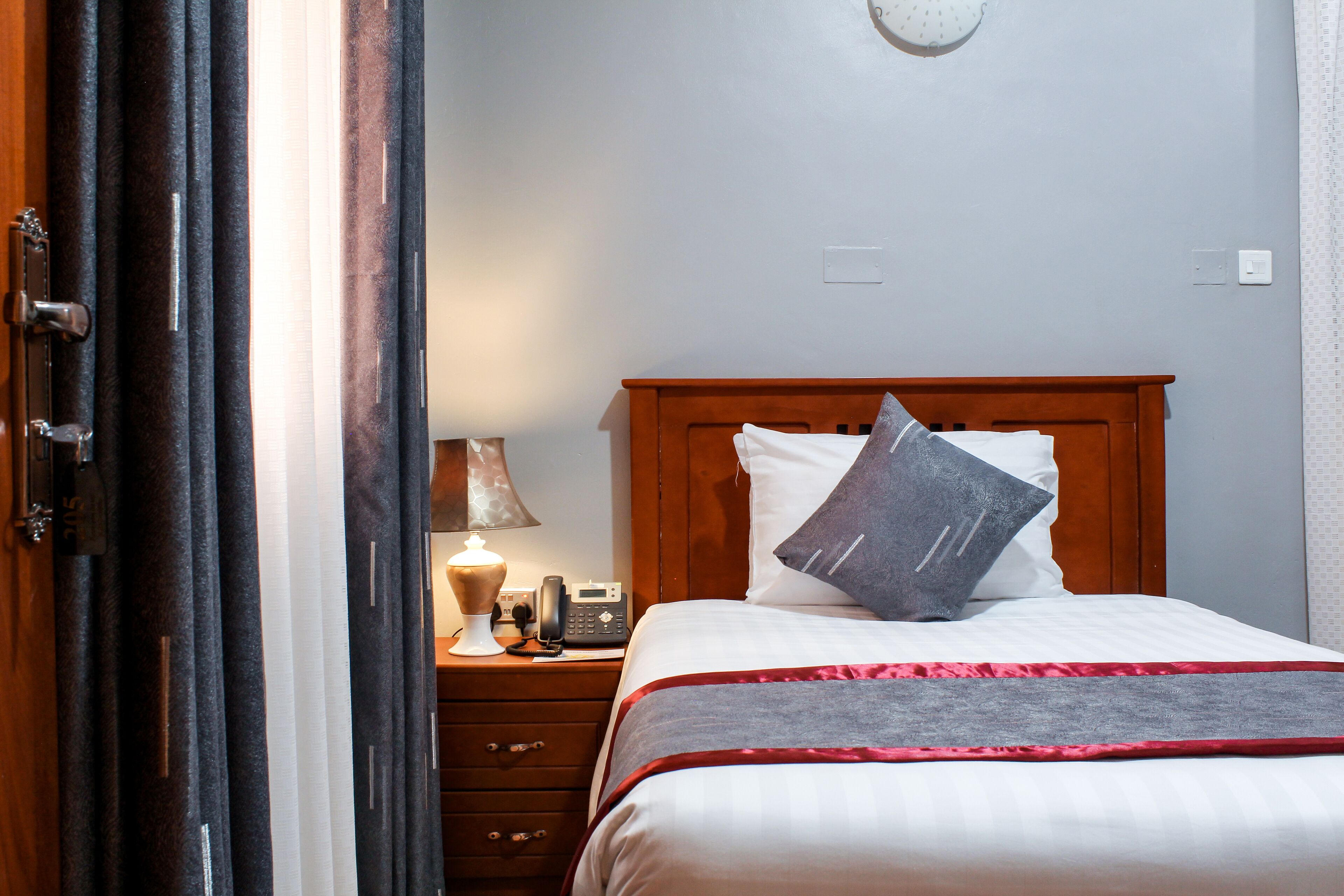 A hotel room with a wooden headboard, a bed with white bedding and a grey throw, a bedside table with a lamp, a telephone, and a wall clock.