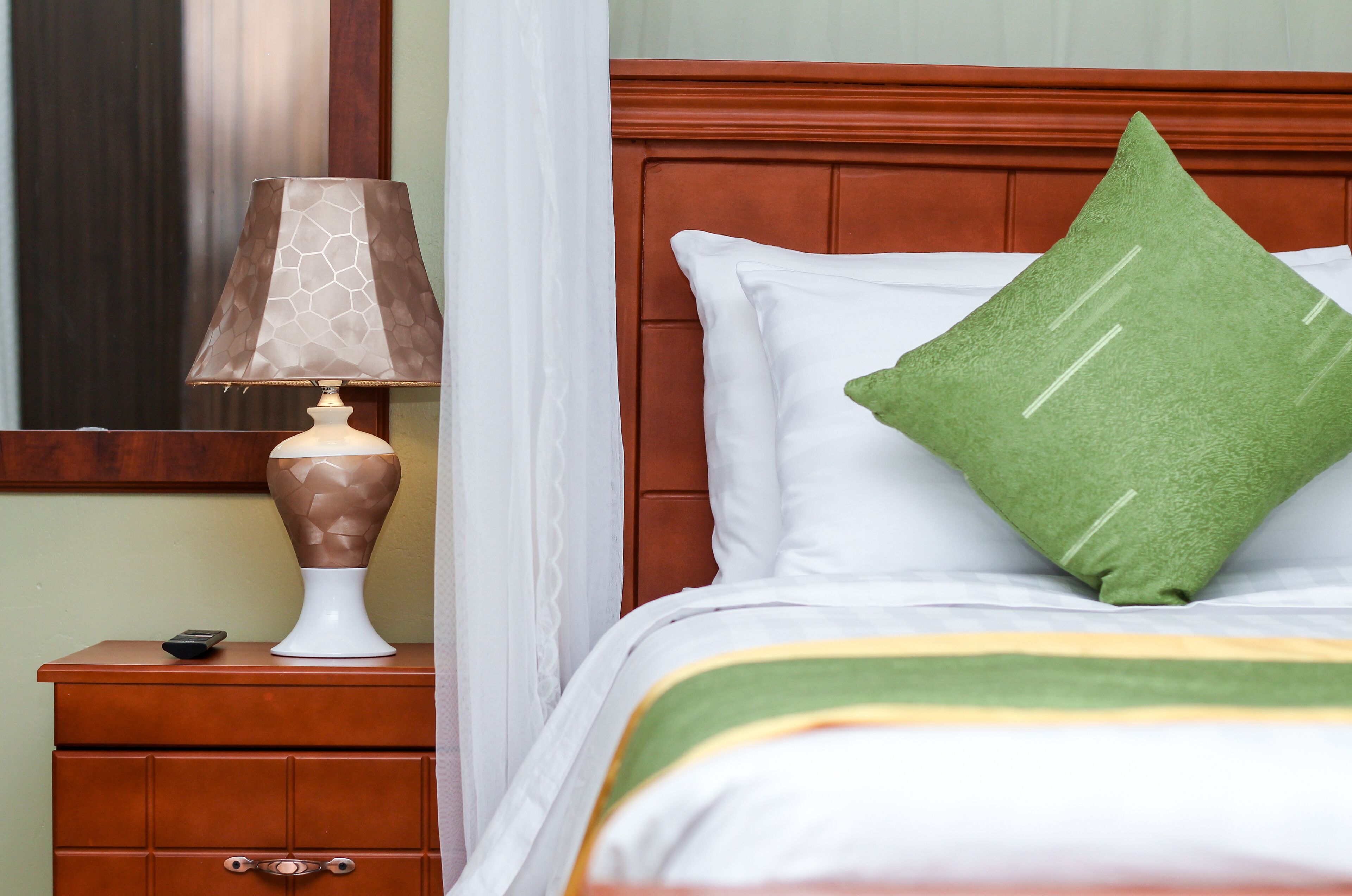 A neatly made bed with a green pillow and a white pillow, a bedside lamp on a wooden nightstand, and a remote control on the nightstand.