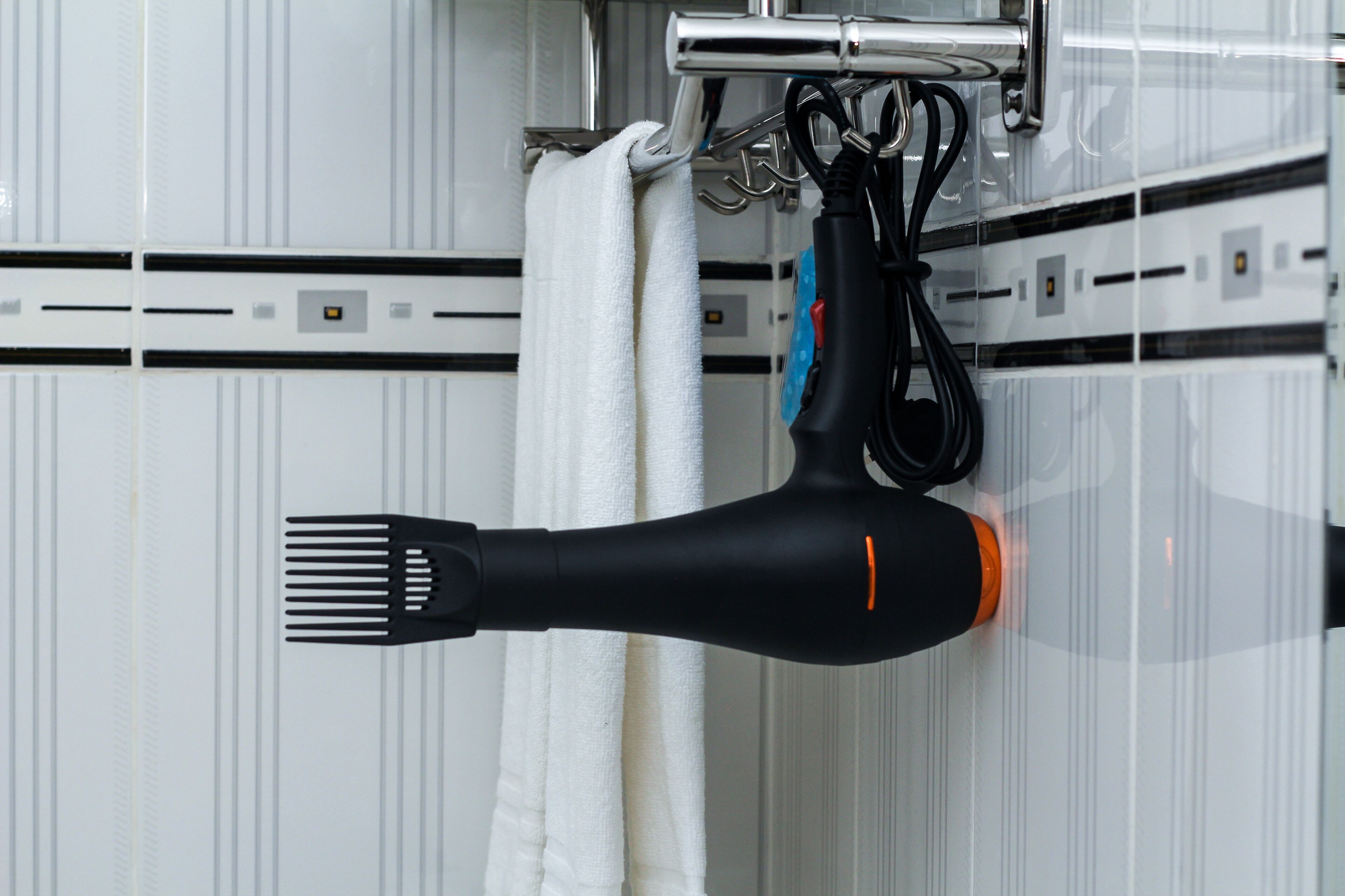 Un sèche-cheveux est suspendu à un porte-serviettes dans une salle de bain dont les murs sont recouverts de carreaux blancs et où une prise électrique est visible.