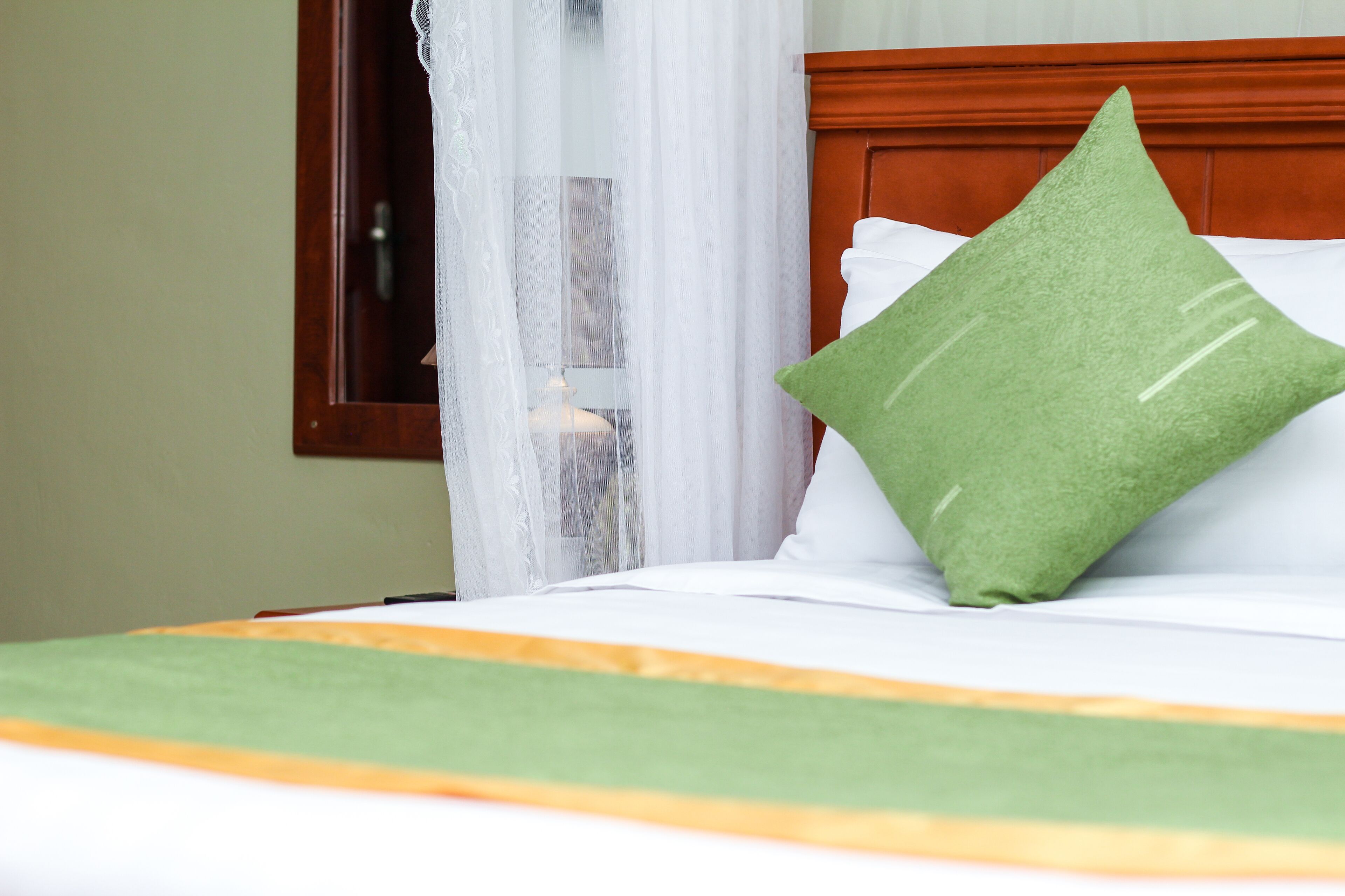 Een net opgemaakt bed met een groen kussen en een wit kussen, een houten hoofdbord en een transparant gordijn dat gedeeltelijk is opengetrokken.