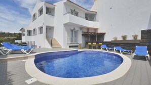 A modern two-story building with a swimming pool, blue lounge chairs, and a staircase leading to the upper floor.