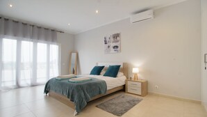 A modern bedroom with a large bed, bedside tables, a nightstand, a window with sheer curtains, and a wall-mounted air conditioner.