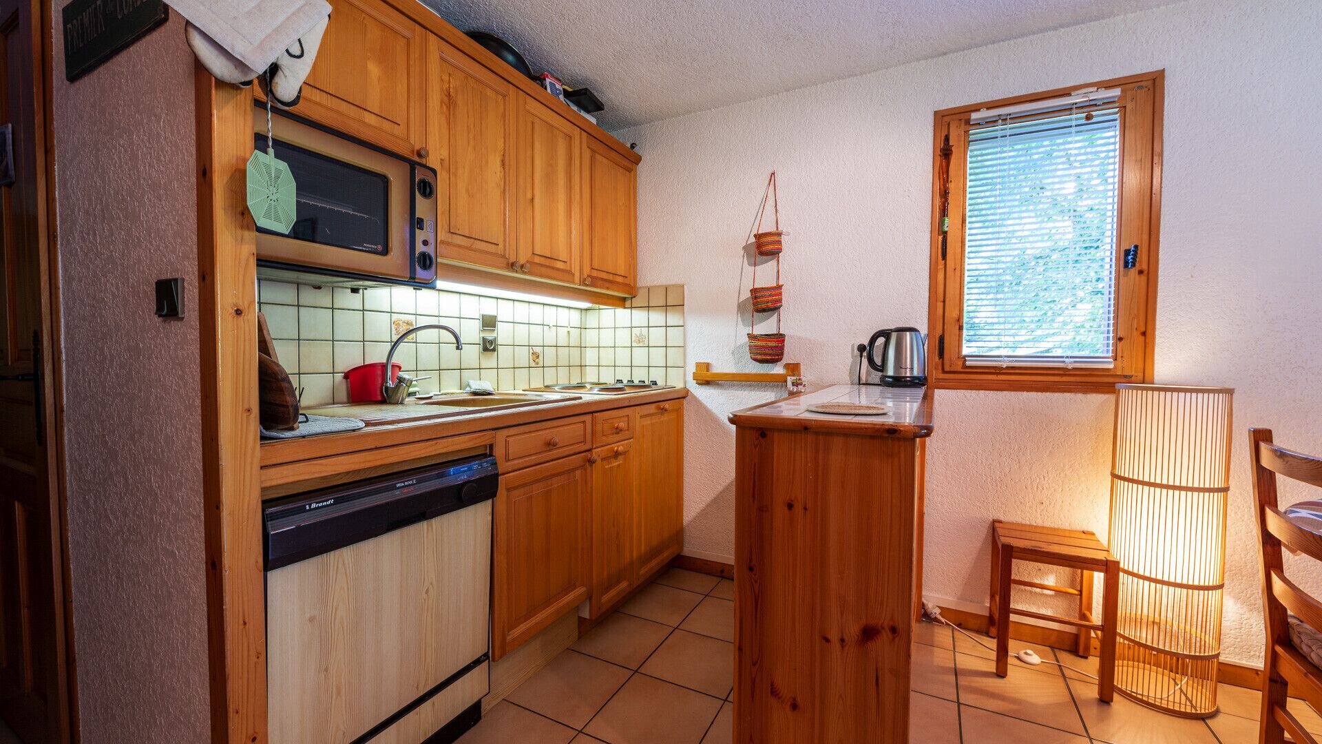 Una cocina compacta con armarios de madera, lavavajillas, microondas y ventana con persianas.