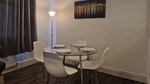 Een eetgedeelte met een ronde glazen tafel, witte stoelen, een vloerlamp en een stadsgezicht schilderij aan de muur.