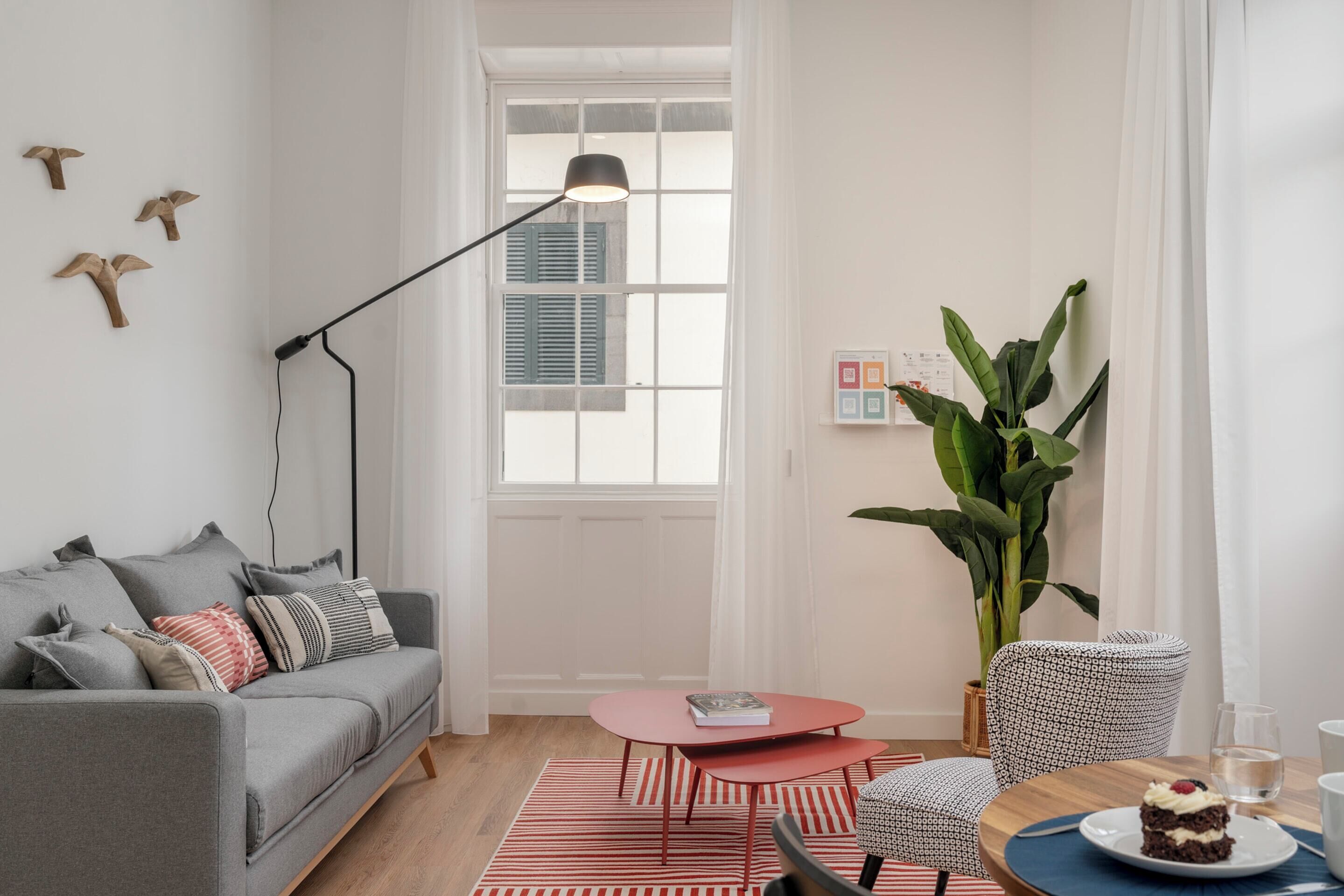 A modern living room with a grey sofa, a red coffee table, and a dining area with a wooden table and chairs.