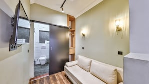 Un baño moderno con suelo de madera, un sofá color beige, un espejo y un inodoro visible a través de una puerta.