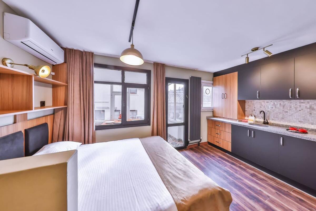A compact hotel room with a bed, a small kitchenette, a window with curtains, and an air conditioning unit.