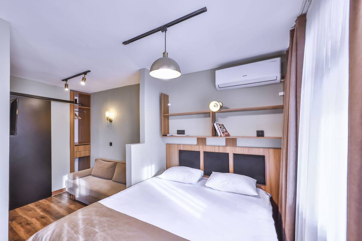A hotel room with a bed, a wardrobe, a shelf with books, a wall-mounted air conditioner, and a hanging light fixture.