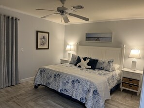 Un dormitorio con una cama, dos mesitas de noche, un ventilador de techo, una lámpara, un cuadro enmarcado y una ventana con cortinas.