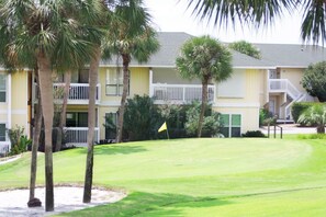 Un campo de golf con una bandera, palmeras y un edificio con balcones.