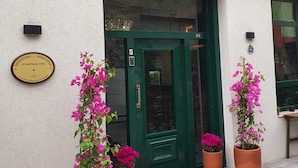 The exterior of the St. 945 Palas Hotel, featuring a green door, a yellow facade, and potted pink flowers at the entrance. - St945 Palas Otel (Izmir)