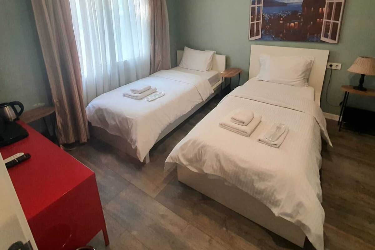 A hotel room with two beds, a red desk, a painting of a cityscape, and a window with curtains.