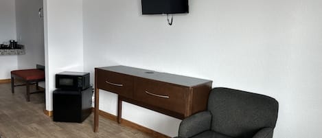 A room with a wooden desk, a chair, a television mounted on the wall, and a microwave on the floor.