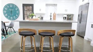 Una cocina con barra, tres taburetes de madera, un reloj grande en la pared y un refrigerador de acero inoxidable.