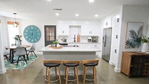 Una cocina moderna con armarios blancos, una isla central con taburetes, un reloj grande y un comedor con mesa y sillas.