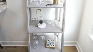 Ein hoher, schmaler, weißer Metallregal mit mehreren Einlegeböden, auf denen sich Glaswaren und ein kleines Dekorationsstück befinden.