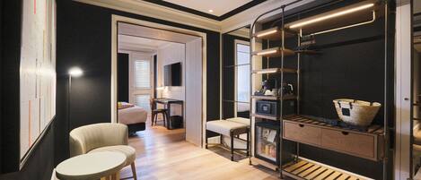 A modern hotel room with a wooden floor, a seating area with a table and chairs, a built-in bench, a wardrobe with shelves, and a hanging rack with baskets.